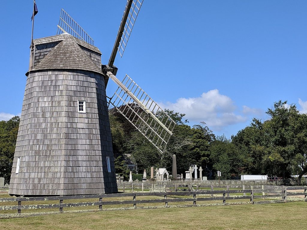 Gardiners Windmill in East Hampton, NY, a historic wooden windmill surrounded by a wooden fence, green grass, and a nearby cemetery under a clear blue sky.