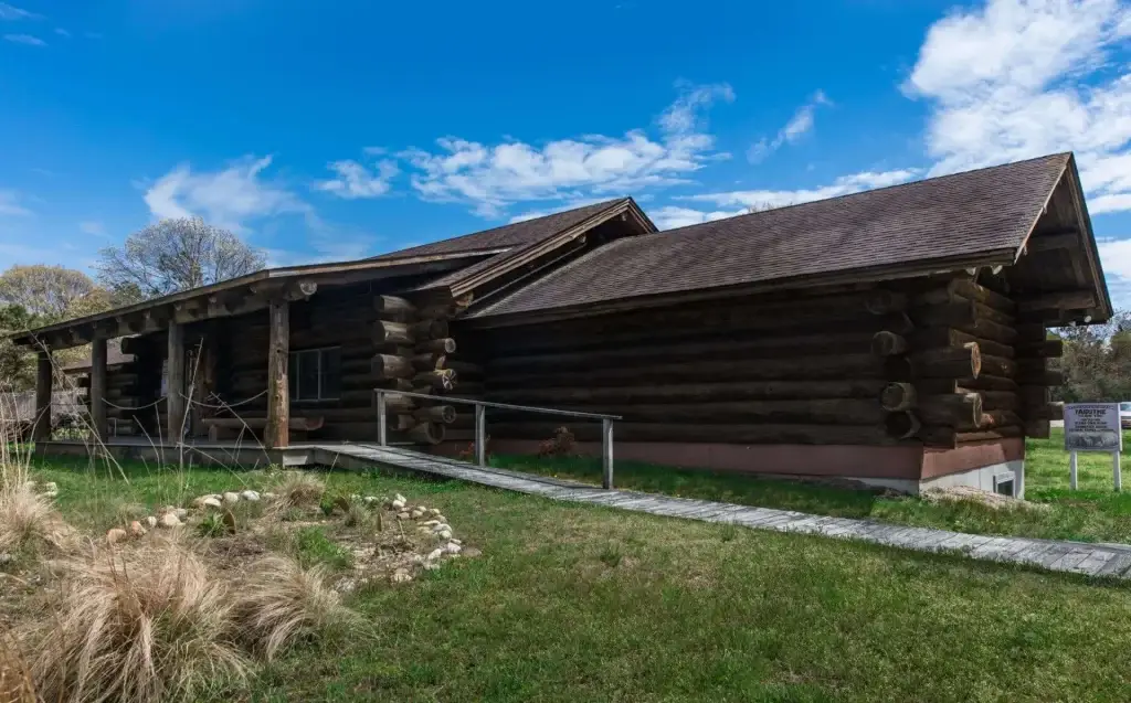 The Shinnecock Nation Cultural Center and Museum in Southampton, NY, a rustic log building surrounded by green grass and a wooden pathway under a bright blue sky.