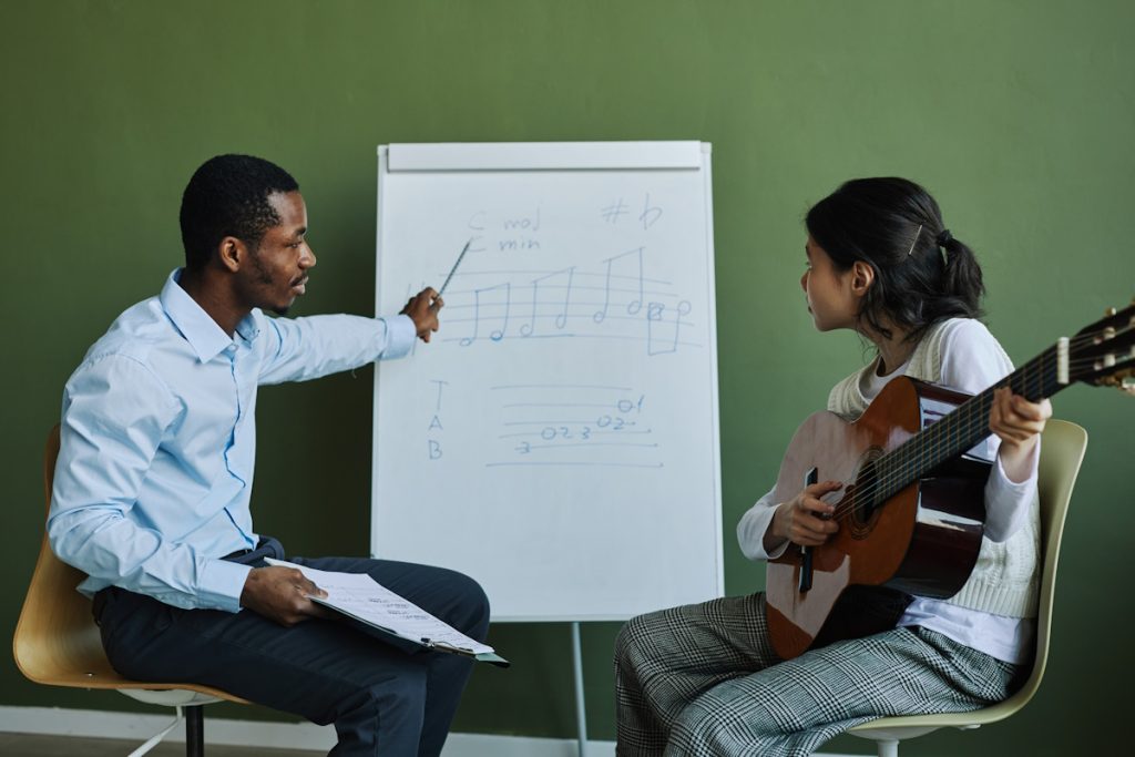 Side view of confident teacher of music explaining musical notes to student with acoustic guitar while pointing at whiteboard