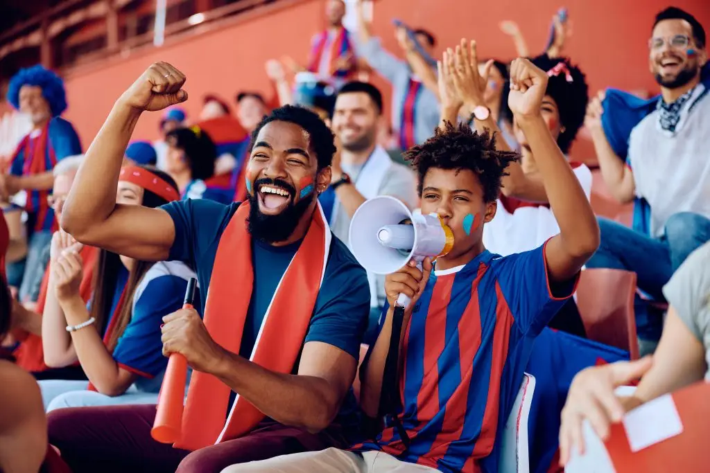 Cheerful fans in a stadium, with a father and son enthusiastically cheering for their team. The father has his fist raised, smiling widely, while the son, holding a megaphone, is dressed in a red and blue striped jersey. The crowd behind them is full of energetic supporters, creating a vibrant and lively atmosphere.