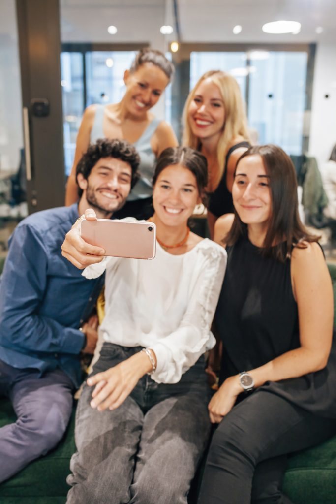 Young creative startup team smiling and taking selfie on mobile phone while gathering together in modern workspace. Blurred group of people.