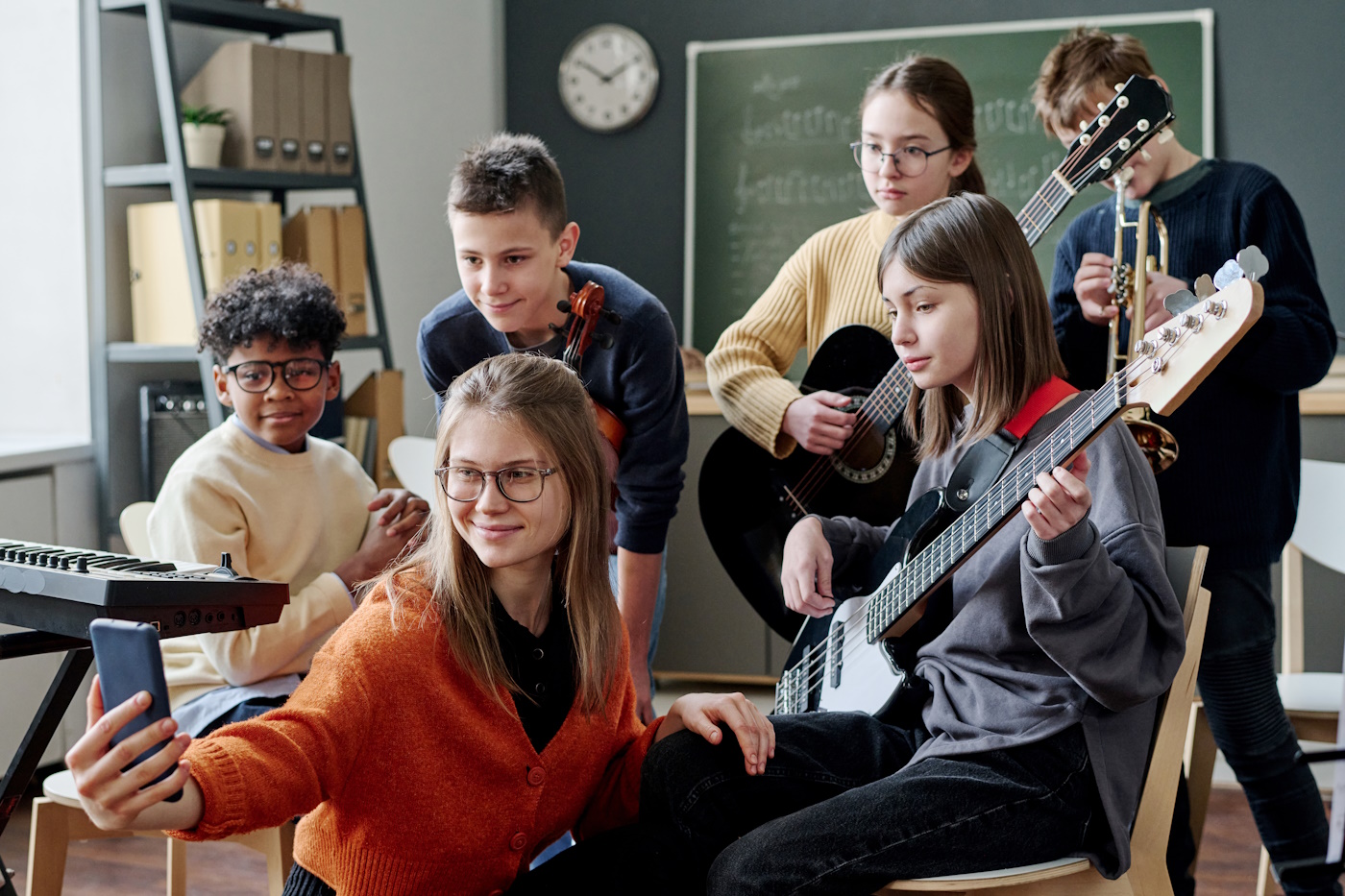 Modern young female music teacher holding smartphone taking selfies with her middle school students in classroom