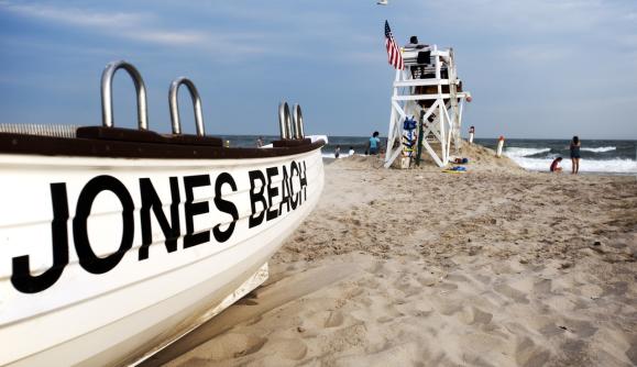 A scenic view of Jones Beach State Park, featuring a lifeguard stand, a lifeboat labeled 'Jones Beach,' and people enjoying the sandy shoreline under a partly cloudy sky.