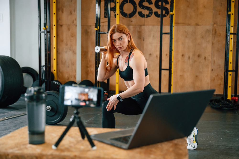 Laptop, bottle and smartphone on tripod is on the table. Beautiful strong woman is in the gym.