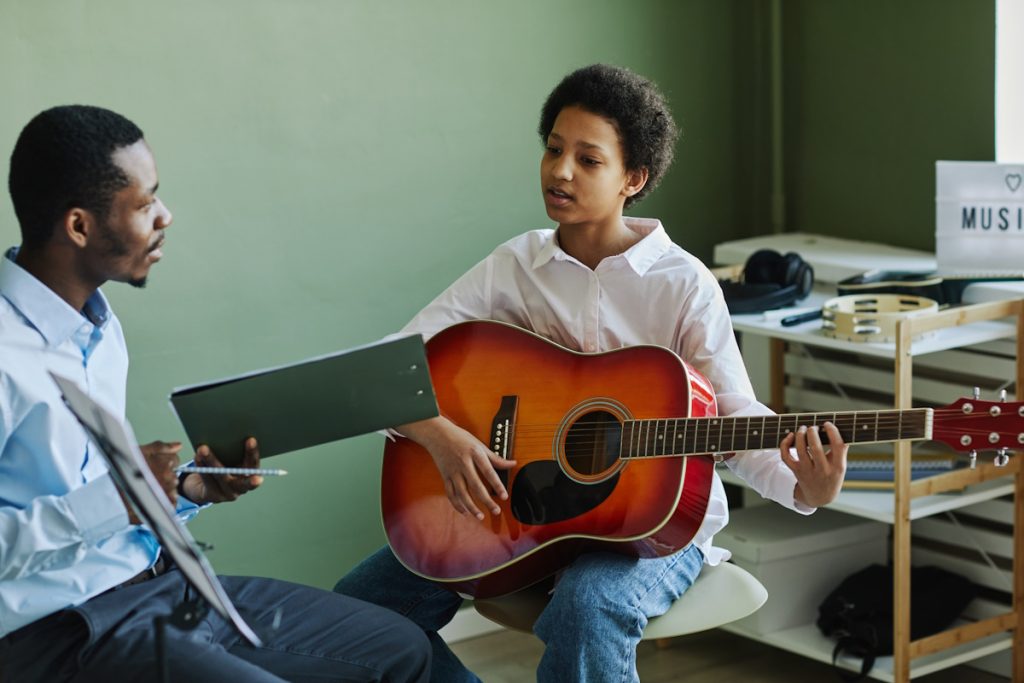 Youthful African American schoolgirl playing acoustic guitar at lesson in music school while sitting in front of teacher consulting her