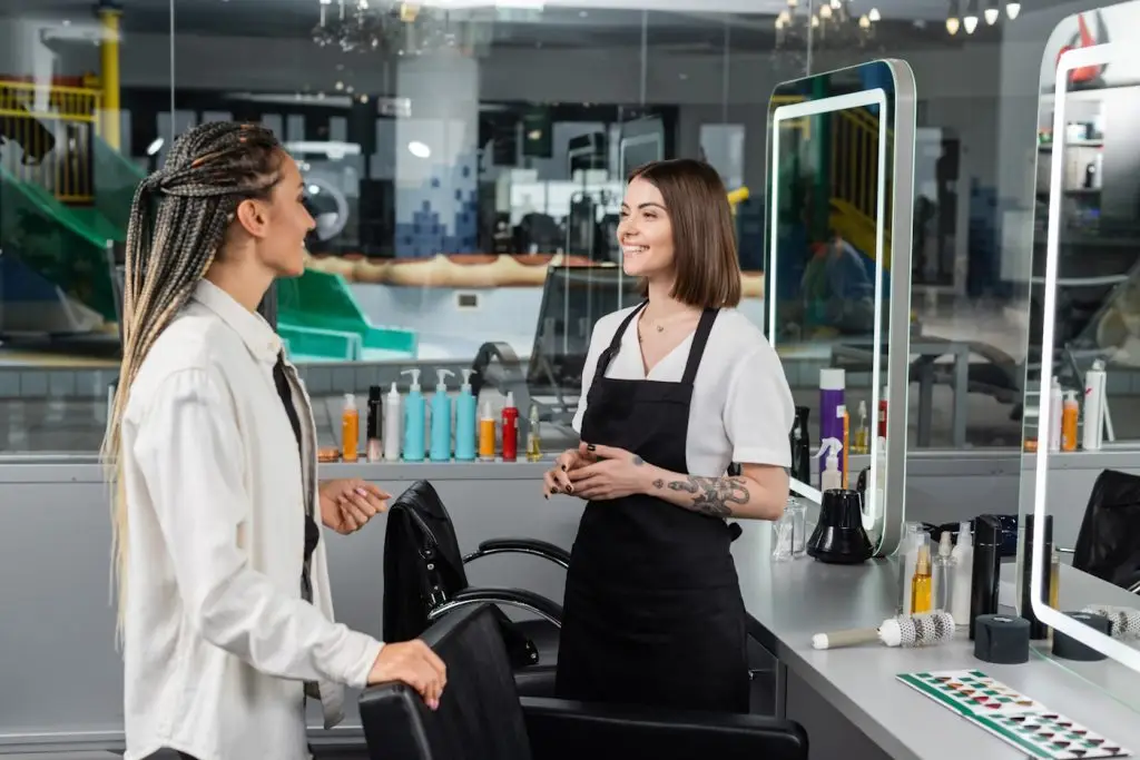 hair professional, happy and tattooed beauty worker in apron welcoming female client with braids in