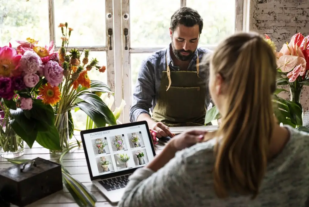 Florist working at a wooden table surrounded by colorful flowers, with a client viewing a flower shop website on a laptop.