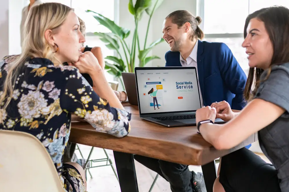 Three colleagues having a discussion around a table with a laptop displaying a social media landing page design.